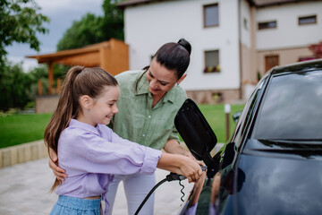 Happy mother showing her little daughter how to charge their electric car.