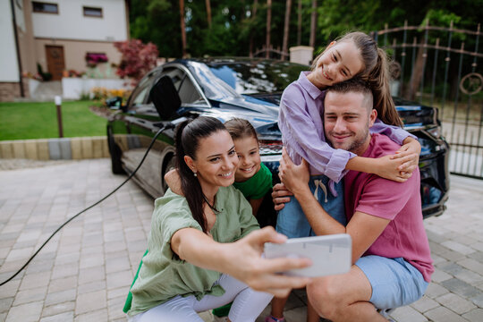 Happy family taking selfie, waiting for electric car charging.