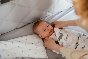 White woman in housecoat swaddling her newborn baby in crib at bedroom