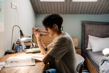 Serious asian guy studying with book while sitting at desk at home