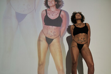 Horizontal medium long low angle shot of young Black woman with tattoos on body wearing black underwear standing with eyes closed in projector light with repeating images of her on background