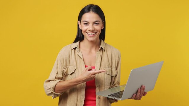 Young latin woman she wear beige shirt pink t-shirt hold in hands typing use work point index finger on laptop pc computer isolated on plain yellow background studio portrait. People lifestyle concept