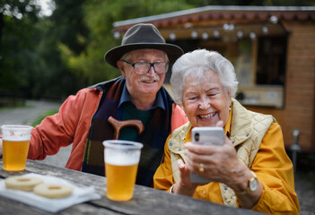 Happy senior couple in forest buffet resting after walk, having beer and looking at smartphone.