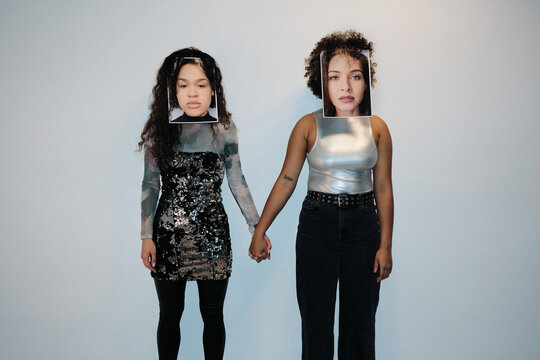 Horizontal medium long studio shot of two young ethnically diverse women covering their own faces with portraits of each other, holding hands, self-identity concept