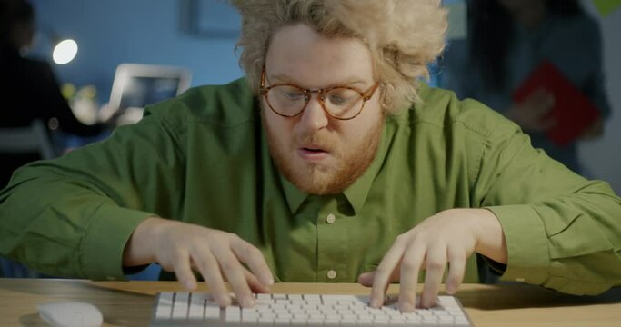 Portrait of comic guy working with computer typing in rush looking at camera with funny face in dark office at night. People and job concept.