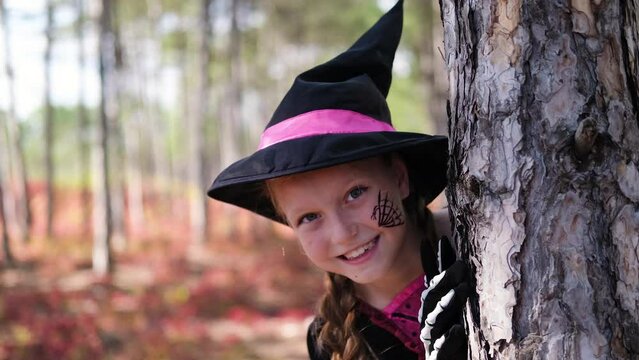 A girl dressed as a witch hides behind a tree in the forest. Halloween child dressed as a witch with face painting
