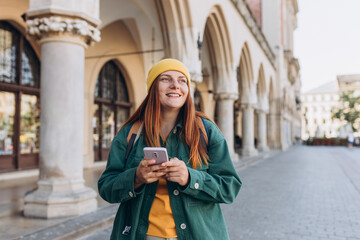 Beautiful stylish woman in hat walking on Market Square in Krakow on autumn day and holding mobile phone. Phone Communication. Urban lifestyle concept. Check social networks, send sms.