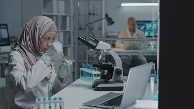 Side view of young female Muslim lab assistant in protective goggles and gloves looking at blue liquid in test tube, sitting at desk in laboratory