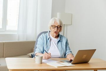 a happy, successful elderly woman is sitting at her desk at home, stylishly dressed in black glasses and happily looking into the camera with a laptop and writing papers on the table
