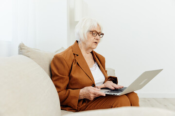 an attentive, pleasant elderly woman is sitting in a bright room at home on a cozy sofa and is holding a video conference for company employees