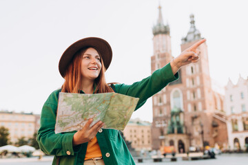 Attractive female tourist is exploring new city. Redhead woman pointing finger and holding a paper map on Market Square in Krakow. Traveling Europe in autumn. St. Marys Basilica. Active lifestyle