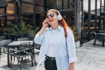 Lovely happy young woman in sunglasess listening to music through wireless earphones on urban background. Music lover enjoying music. Lifestyle concept. Girl walking on city street