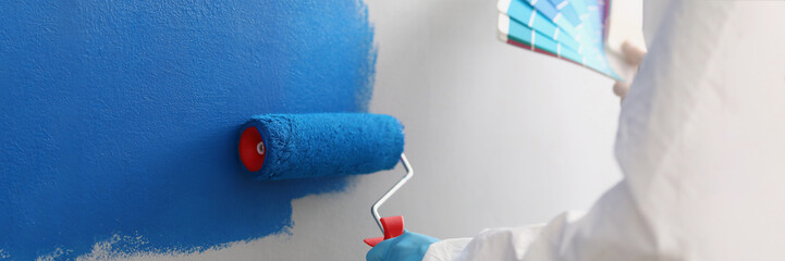 Painter with brush tool covered in blue colour painting wall, worker on site