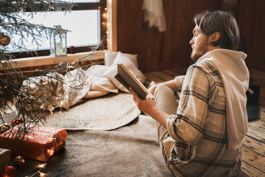 Young stylish man is stoking stove, reading cook. Celebrating new year eve in cozy wooden country house in winter forest. Gifts,family holiday.Romantic weekend Christmas tree with gold red xmas balls
