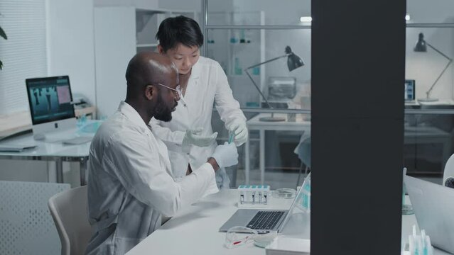 Medium side-view of Black male scientist in white gown typing on laptop computer, sitting at desk in modern laboratory, his female Asian colleague bringing him sample in tube