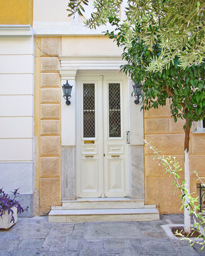 A classic design house front entrance, with white painted wooden doors, ocher walls and a lemon tree by the sidewalk. Plaka, Athens, Greece.