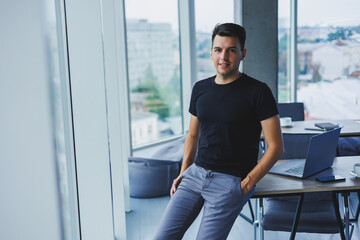 Portrait of a smiling young man in a black t-shirt against the background of a modern office. Stylish guy in casual clothes