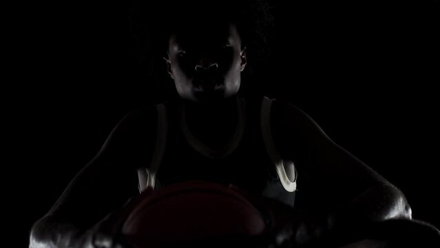 Sidelit basketball player silhouette sitting on a bench holding a ball. Concentrated african american sports man sidelit against black background.