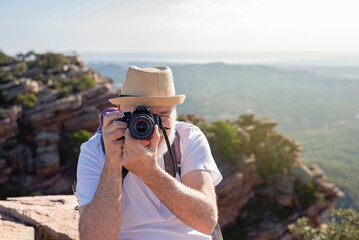 front view of an older hiker sitting on the summit taking a picture with his camera.