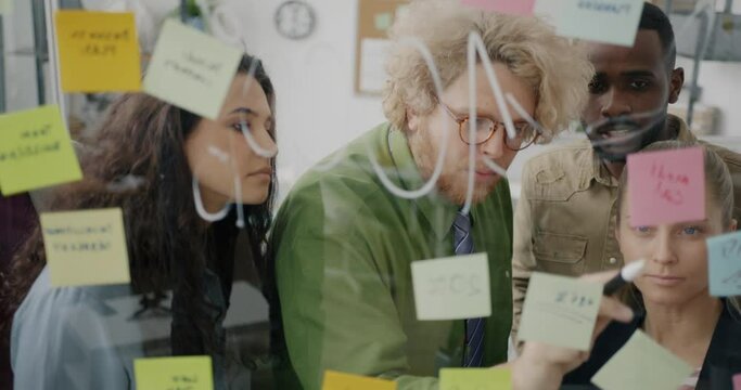 Joyful businesspeople working with sticky notes on glass board talking and writing thinking about business development. Creative team and job concept.