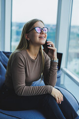 Young smiling woman wearing glasses in casual clothes sitting in a chair by the window and talking on the phone during a video call.