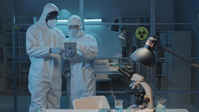 Medium long tracking left of female Asian and male African American doctors in protective suits standing in laboratory, using tablet computer, talking