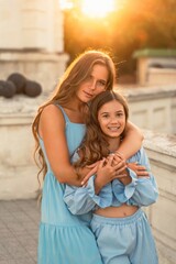 Portrait of mother and daughter in blue dresses with flowing long hair against the backdrop of sunset. The woman hugs and presses the girl to her. They are looking at the camera.