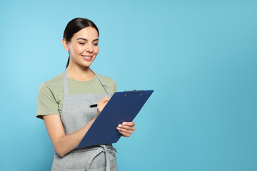 Young woman in grey apron with clipboard on light blue background, space for text