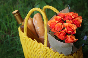Fototapeta na wymiar Yellow wicker bag with beautiful roses, bottle of wine and baguettes on green grass outdoors, closeup
