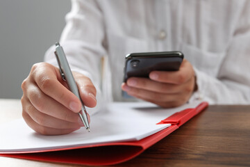 Woman with smartphone writing on sheet of paper in red folder at wooden table in office, closeup