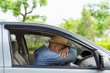 Closeup portrait sleepy tired fatigued exhausted young man wear hat driving his car in street traffic after long hour trip side view. Handsome Young male sleeping in a car, resting head on wheel.