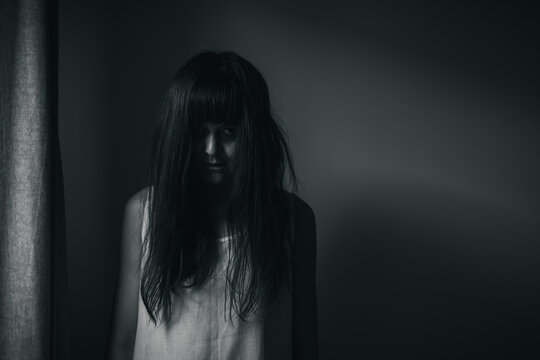 Female ghost in white dress in room. A young scary girl in an old white dress staring in to the camera ferociously with dark background. Asian woman make up ghost face at house. Scary horror concept.