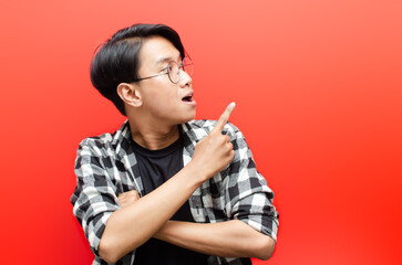 asian young man with glasses shocked happily pointing up to copy space isolated over red background. billboard advertisment model concept.