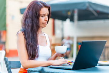 Beautiful young woman in white t-shirt with long hair is working on laptop and smiling while sitting outdoors in cafe.Young female drinks coffee.Female freelancer working on laptop in an outdoor cafe.