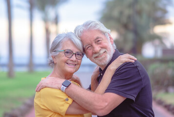 Portrait of happy lovely senior couple embracing in outdoor public park at sunset. Two caucasian elderly people looking at camera smiling enjoying good time together