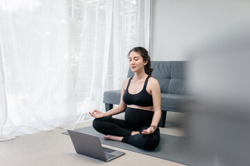Beautiful pregnant asian woman is meditating in a lotus position in front of her laptop for the online yoga practicing in the living room to keep her prenatal healthy. Concept of Healthy Pregnancy.