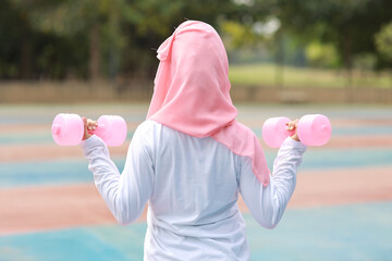 Rear view athletic young asian woman in muslim sportswear standing and lifting dumbbells outdoor for morning exercise. Active girl work out on biceps exercise with green tree background. Sport concept