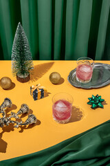 Vivid color retro futuristic Christmas dinner table setting with a tree, gift, yellow bow, red ice cold cocktails in crystal glass with emerald green curtain background. New Year's eve party aesthetic