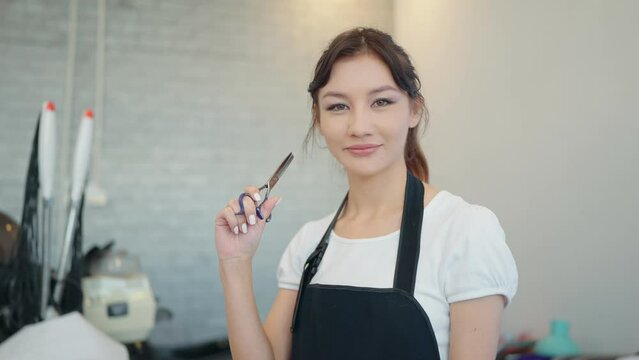 Portrait of smiling beautiful woman hairdresser with black apron at camera while holding scissors at hair salon
