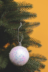 White holographic Christmas decoration hanging on a fir tree. New Year bauble in front of yellow background.