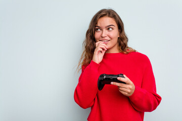 Young caucasian gamer woman holding a game controller isolated on blue background relaxed thinking about something looking at a copy space. © Asier