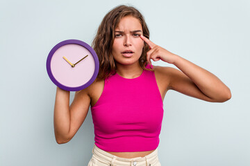 Young caucasian woman holding a clock isolated on blue background showing a disappointment gesture with forefinger.