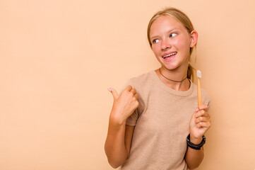 Caucasian teen girl brushing teeth isolated on beige background points with thumb finger away, laughing and carefree.