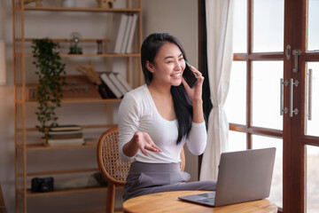 Obraz premium Asian woman sitting at home working on her laptop and talks on the phone comfortably and refreshed on a new day.