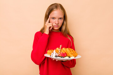 Little caucasian girl holding a waffles isolated on beige background pointing temple with finger, thinking, focused on a task.