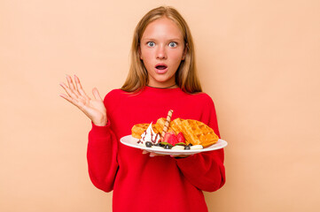 Little caucasian girl holding a waffles isolated on beige background surprised and shocked.