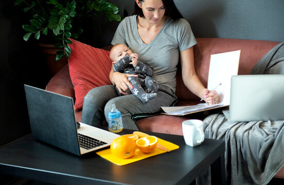 A Young Mom Is Holding A Baby And Working On A Laptop And Documents At Home Office On The Couch