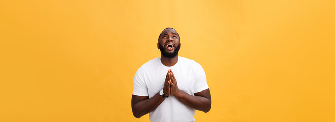 Studio portrait of young African American man in white shirt, holding hands in prayer, looking at the camera with thoughtful skeptical expression on his face, suspecting of something. Body language.