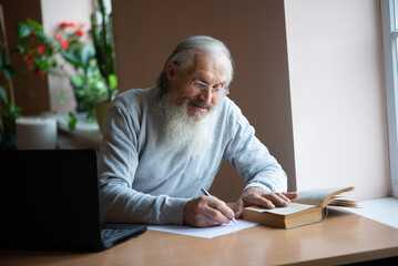 Bearded old man with laptop and book sitting at table and write notes... © Zelma