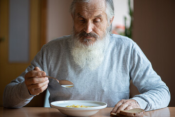 Old sad man with a long gray beard sitting by the table and eating soup and bread